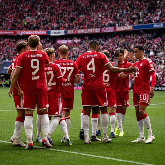 Des joueurs de football du FC Bayern Munich, en maillots rouges, se réunissent sur le terrain, célébrant joyeusement en se serrant la main et en se tapant dans la main. En arrière-plan, des supporters remplissent les gradins.