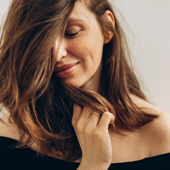 A woman with long, flowing hair gently brushes her fingers through it, smiling softly. She is wearing an off-shoulder top and has a serene expression, conveying a sense of calm and contentment. The background is simple and unobtrusive.