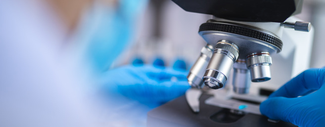 Un scientifique en blouse de laboratoire utilise un microscope, avec des gants en latex bleus, pour examiner un échantillon. Le microscope est bien détaillé, tandis que l'arrière-plan est flou, mettant l'accent sur l'équipement de laboratoire.