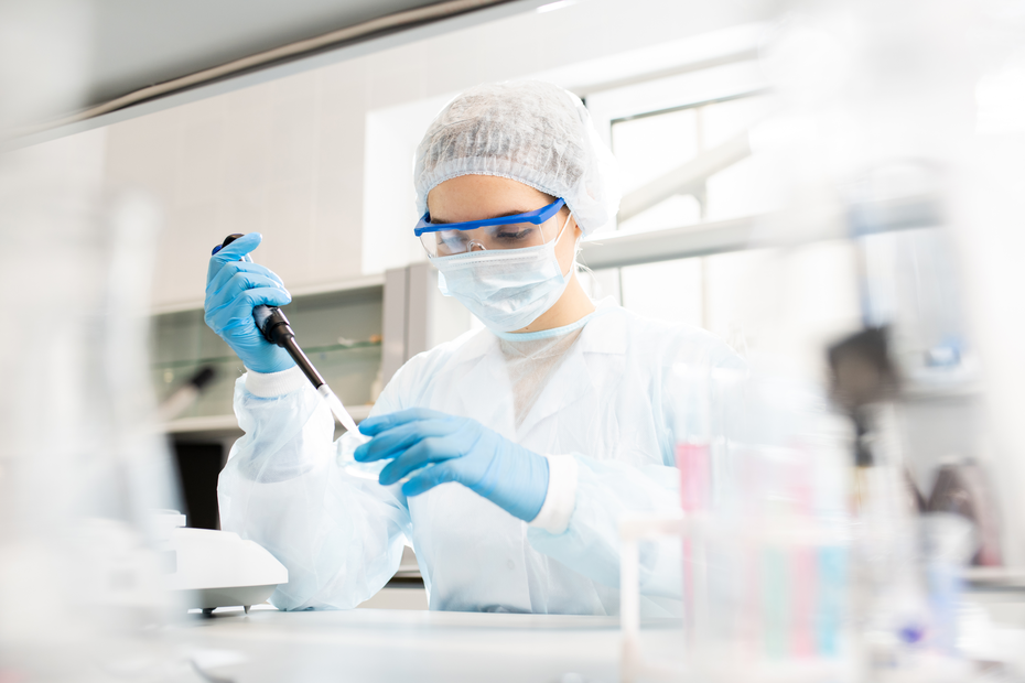 Une scientifique en blouse blanche, gantée et portant des lunettes de protection et un masque, utilise une pipette dans un laboratoire. Elle se concentre sur son travail, entourée d'instruments et de récipients de laboratoire.