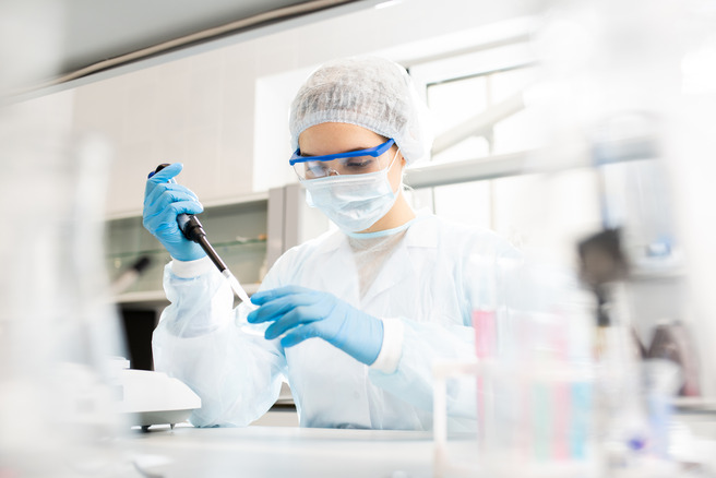 Une scientifique en blouse blanche, gantée et portant des lunettes de protection et un masque, utilise une pipette dans un laboratoire. Elle se concentre sur son travail, entourée d'instruments et de récipients de laboratoire.
