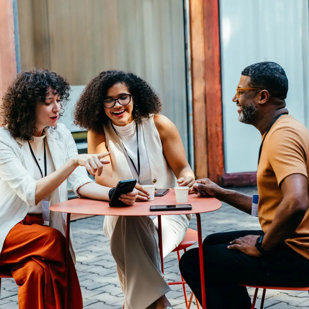 Trois personnes assises à une table extérieure, discutant et riant ensemble. Deux femmes avec des cheveux bouclés et un homme avec des lunettes échangent des sourires. Des téléphones et des boissons sont visibles sur la table.
