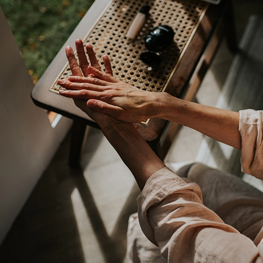 A person's hands are shown applying lotion to one hand while resting on a textured surface. The scene conveys a sense of self-care and relaxation, with soft natural lighting enhancing the calming atmosphere.