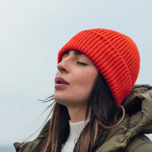 A woman with long dark hair is wearing an orange knitted beanie and a warm jacket. She has her eyes closed and appears to be enjoying the fresh air, with a serene expression on her face against a soft, cloudy background.