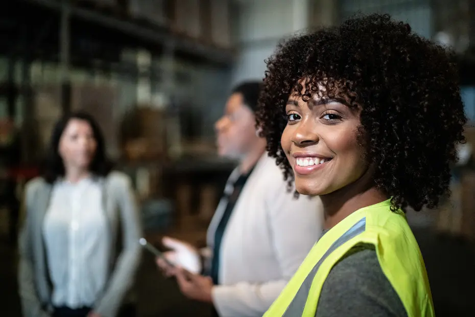 Trois femmes sont présentes dans un environnement de travail. La femme au premier plan, souriante, porte un gilet de sécurité jaune. En arrière-plan, deux autres femmes discutent. L'ambiance est industrielle, avec des étagères chargées de matériel.