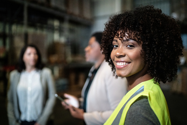 Trois femmes sont présentes dans un environnement de travail. La femme au premier plan, souriante, porte un gilet de sécurité jaune. En arrière-plan, deux autres femmes discutent. L'ambiance est industrielle, avec des étagères chargées de matériel.