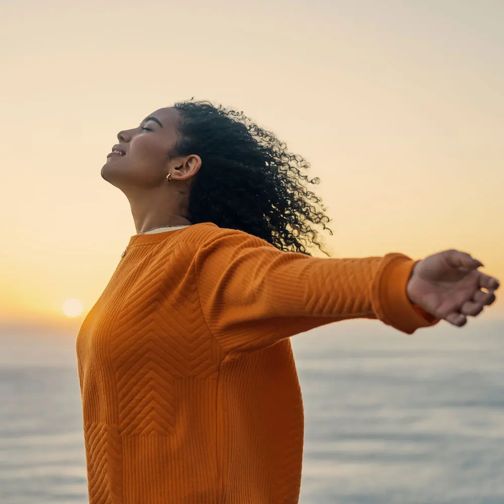 Une femme avec des cheveux bouclés se tient face à l'horizon, les bras grands ouverts, comme pour accueillir le lever du soleil. Elle porte un pull orange et semble respirer profondément, exprimant la joie et la sérénité.