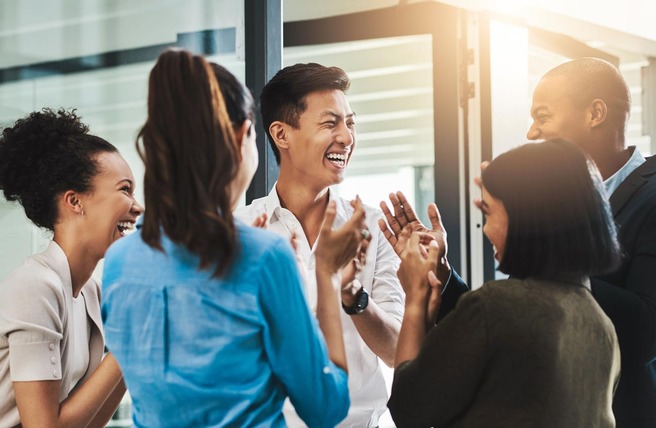 Un groupe de cinq personnes rit et applaudit joyeusement dans un espace de bureau lumineux. Elles semblent engagées dans une conversation conviviale, exprimant de la joie et de l'enthousiasme.