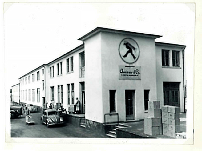 Bâtiment d'une entreprise avec une enseigne circulaire représentant un homme en marche. Des boîtes empilées sont visibles à l'extérieur, et plusieurs personnes et voitures anciennes se trouvent devant l'entrée. L'image est en noir et blanc.