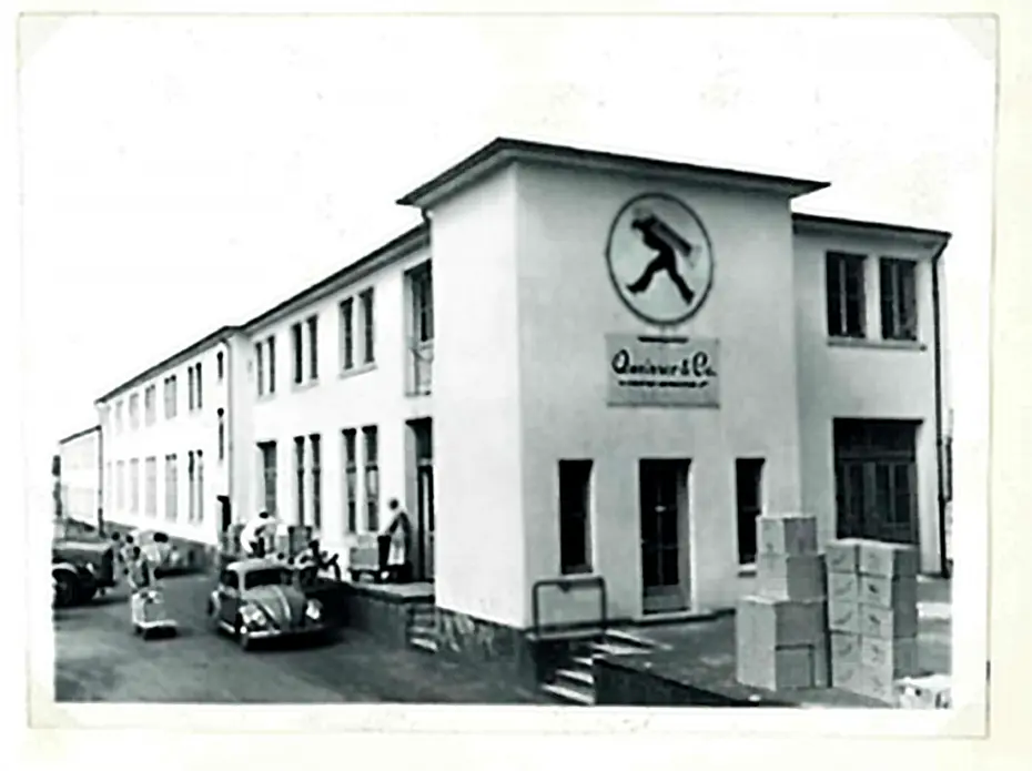 Bâtiment d'une entreprise avec une enseigne circulaire représentant un homme en marche. Des boîtes empilées sont visibles à l'extérieur, et plusieurs personnes et voitures anciennes se trouvent devant l'entrée. L'image est en noir et blanc.