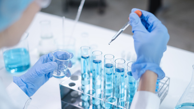 Un scientifique en blouse blanche et gants bleus utilise une pipette pour ajouter un liquide dans des tubes à essai remplis de solution bleue. Un bécher est tenu dans l'autre main, sur une table de laboratoire avec divers équipements.
