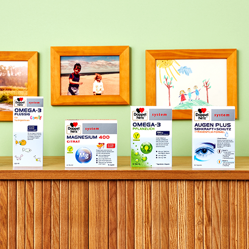 Sur un meuble en bois, plusieurs boîtes de suppléments santé sont alignées : Omega-3, Magnésium 400 Citrate, et Augen Plus. En arrière-plan, des photographies de famille et un dessin enfantin décorent un mur de couleur claire.