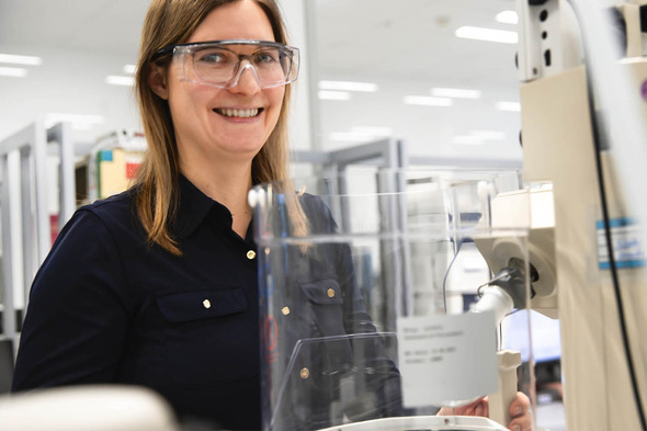 Femme souriante portant des lunettes de sécurité dans un laboratoire. Elle est devant un équipement scientifique, avec un environnement de travail moderne et organisé en arrière-plan.