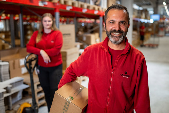 Deux employés d'un entrepôt, l'un souriant et tenant une boîte, l'autre en arrière-plan, détendue près d'un transpalette. Tous deux portent des vêtements rouges dans un environnement de travail rempli de boîtes.