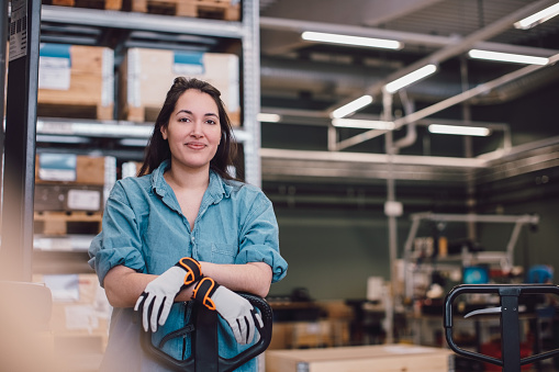 Une femme souriante portant un chapeau et des gants de travail se tient devant des palettes en bois dans un environnement d'entrepôt. Elle pose ses mains sur un transpalette, avec des étagères chargées de marchandises en arrière-plan.